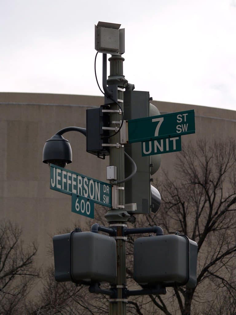 A police surveillance camera and antenna mounted on a streetlight pole above a city intersection.